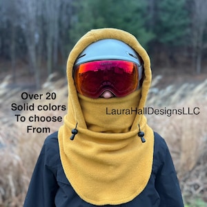 May include: A person wearing a mustard yellow fleece hood and a gray ski helmet with red mirrored goggles. The hood has black toggles and is worn over a black jacket. Text on the image reads "Over 20 Solid colors To choose From" and "LauraHallDesignsLLC".