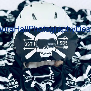 May include: A white ski helmet with a black skull and crossbones design. The skull is wearing goggles and the crossbones are ski poles. The text "GST EST 2010 SD5" is printed on the helmet.