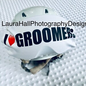 Peut inclure: Casque de ski blanc avec une housse de casque de ski blanche et noire qui dit "I ♥ Groomers" en texte noir avec un cœur rouge.
