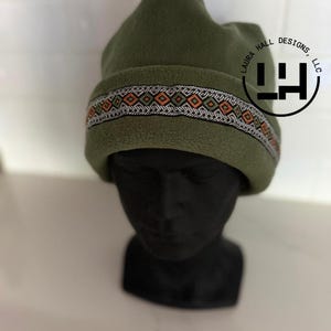 May include: An olive green fleece beanie featuring a folded cuff with a decorative woven band. The band displays a repeating geometric pattern in orange, white, and black. A circular logo with the text LAURA HALL DESIGNS, LLC is visible.