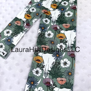 May include: A green fabric with a white cow skull and floral pattern. The flowers are in shades of pink, orange, yellow, and white. The fabric has the text "LauraHallDesignsLLC" printed on it.