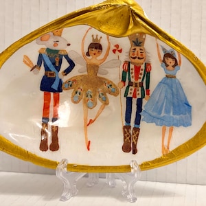 Peut inclure: Un plat décoratif en coquillage avec un bord doré et une scène peinte de personnages du ballet Casse-Noisette. Les personnages comprennent une souris, une ballerine, un casse-noisette et une fille en robe bleue. Le plat est soutenu par un support transparent.