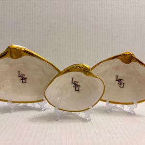 May include: Three white clam shell dishes with gold rims and the letters "LSU" in purple. The shells are displayed on clear plastic stands. The shells vary in size, with the largest on the left and the smallest in the center.