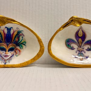 May include: Two decorative seashell dishes with gold-painted rims. One features a colorful Mardi Gras mask design, and the other displays a purple fleur-de-lis. The interior of each shell is white.