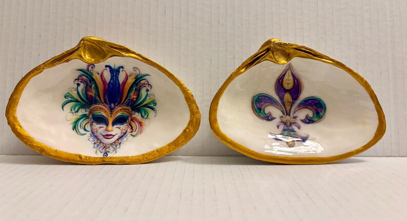 May include: Two decorative seashell dishes with gold-painted rims. One features a colorful Mardi Gras mask design, and the other displays a purple fleur-de-lis. The interior of each shell is white.