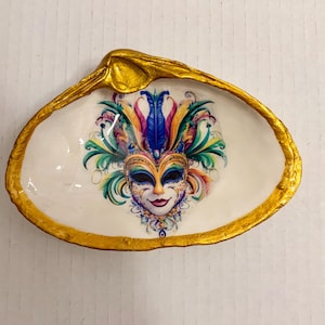 May include: A decorative seashell trinket dish with a gold-painted rim. The interior features a colorful mask design with blue, green, and purple accents. The dish is white and has a glossy finish, perfect for holding small items.