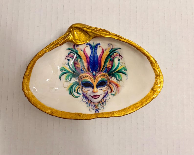 May include: A decorative seashell trinket dish with a gold-painted rim. The interior features a colorful mask design with blue, green, and purple accents. The dish is white and has a glossy finish, perfect for holding small items.