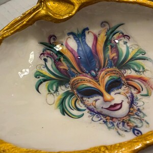 May include: A decorative, oval-shaped trinket dish with a gold-painted rim. The dish features a colorful Mardi Gras mask design in the center, with a white background. The mask has feathers in shades of blue, green, yellow, and purple.