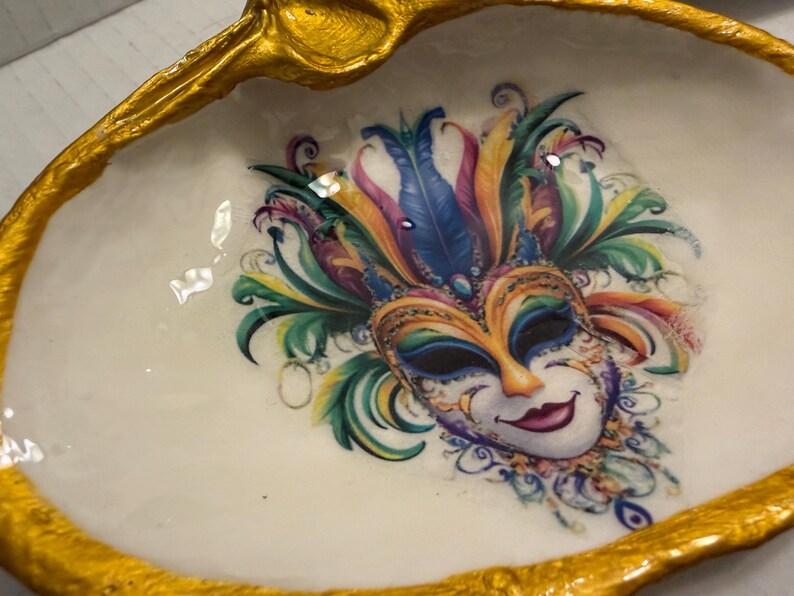 May include: A decorative, oval-shaped trinket dish with a gold-painted rim. The dish features a colorful Mardi Gras mask design in the center, with a white background. The mask has feathers in shades of blue, green, yellow, and purple.