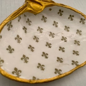 May include: A decorative seashell-shaped dish with a gold-painted rim. The interior is white with a repeating pattern of gold fleur-de-lis symbols. This trinket dish is ideal for holding small jewelry items.