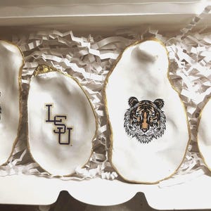 Puede incluir: Cuatro conchas de ostra blancas con bordes dorados, dispuestas en una caja con papel triturado. Dos conchas presentan un gráfico de cabeza de tigre, y dos muestran las letras "LSU" en negro y dorado. Un artículo decorativo.
