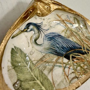 May include: A decorative seashell dish with a gold-painted rim. The interior features a detailed illustration of a blue heron amidst reeds and foliage. The artwork is set against a white background.