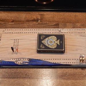 Op de afbeelding: Een houten cribbagebord met een blauwe accentkleur en een kaartspel. Het bord heeft gaten voor pinnen en een kaartspel met een gouden schoppenontwerp. Inclusief pinnen in verschillende kleuren.