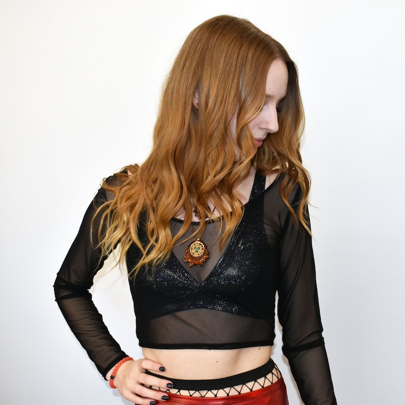 K&ouml;nnte beinhalten: Eine Frau tr&auml;gt ein schwarzes Netz-Crop-Top mit langen &Auml;rmeln und einer glitzernden Textur. Sie tr&auml;gt eine braune und goldene Halskette mit Anh&auml;nger und einen rot-schwarzen Rock.