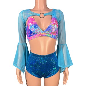 May include: A blue mesh crop top with long bell sleeves and a keyhole neckline. The top is worn over a colorful abstract print crop top. The model is also wearing high-waisted blue shorts with a sparkly finish.