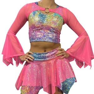 May include: A vibrant, two-piece dance costume set. The top features a keyhole design with a gold ring and long, sheer pink sleeves. The set includes a matching skirt and shorts, all in a shimmering, iridescent fabric with pink and blue accents.