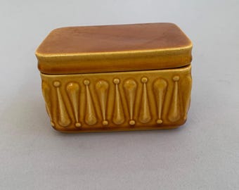May include: A rectangular ceramic butter dish with a smooth, flat, dark amber lid and a mustard yellow base. The base features a repeating vertical pattern of raised, teardrop-shaped motifs with small circular accents along the bottom edge.