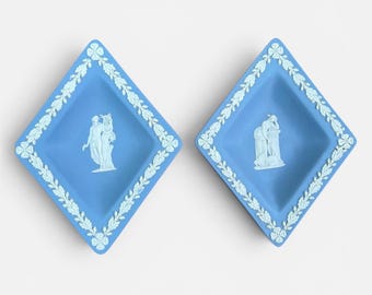 May include: Two blue diamond-shaped dishes with white relief designs. Each dish features a central classical figure scene and a white floral border. The dishes are displayed on a white background.