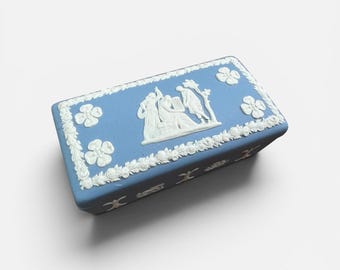 May include: A rectangular blue trinket box with white floral and relief designs. The lid features a white relief scene of figures and a tree. The box is decorated with white flowers and a floral border.