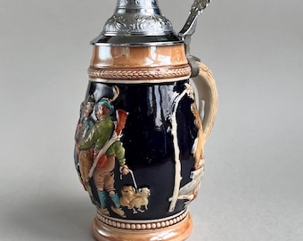 May include: A ceramic beer stein with a dark blue body, featuring a relief scene of hunters with a dog. The stein has a silver-coloured metal lid and handle, and a tan base and rim. The handle is beige and the stein is decorated with a hunting scene.