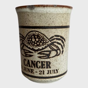 Op de afbeelding: Een keramische mok met een gespikkelde beige afwerking, met een zwarte krabillustratie en het woord "CANCER" met de data "JUNI - 21 JULI". De rand is bruin en de mok heeft twee zwarte banden.