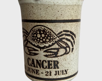 May include: A ceramic mug with a speckled beige finish, featuring a black crab illustration and the word "CANCER" with the dates "JUNE - 21 JULY". The rim is brown, and the mug has two black bands.