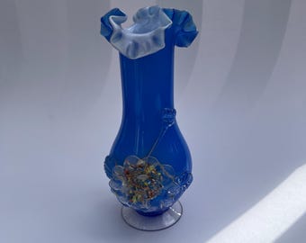 May include: A vibrant blue glass vase featuring a ruffled white rim and a bulbous body. The exterior is decorated with a clear glass floral appliqué containing multicolored confetti-like flecks, set against a smooth, translucent blue surface.