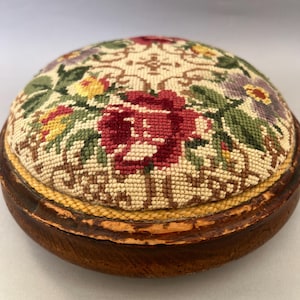 Puede incluir: Un acerico redondo con un patrón floral de punto de aguja con flores rojas, amarillas y moradas sobre un fondo crema, colocado dentro de una base circular de madera marrón oscuro con un borde trenzado amarillo alrededor de la tela.