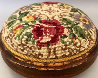 May include: A round pincushion featuring a needlepoint floral pattern with red, yellow, and purple flowers on a cream background, set within a circular dark brown wooden base with a yellow braided trim bordering the fabric insert.