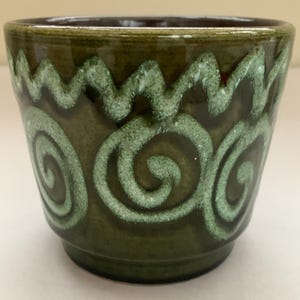 Puede incluir: Una maceta de cerámica verde con un diseño de espiral blanco. La maceta tiene un borde marrón y un diseño ondulado alrededor de la parte superior.