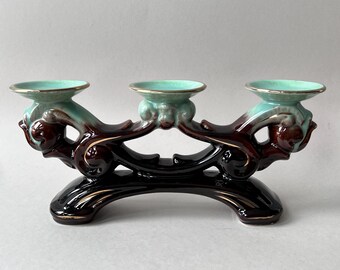 May include: A three-cup candle holder with a decorative design. The ceramic holder features a black base, brown and teal accents, and gold trim. The candle holders are teal with gold rims.