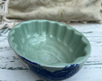 May include: A light green ceramic serving dish with a scalloped edge and a blue patterned rim. The dish has a smooth, glossy finish and a shallow, oval shape. The blue pattern appears to be a stylized landscape.