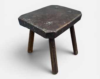 May include: A small, rustic wooden stool with a rectangular seat and three legs. The stool is made of dark brown wood with visible grain and nail heads. The legs are slightly tapered and angled outward for stability. A vintage, handcrafted piece.
