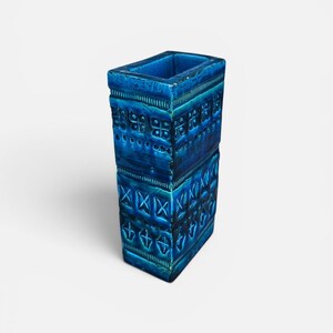 May include: A rectangular, ceramic vase in a vibrant shade of blue. The vase features a textured surface with geometric patterns, including squares, crosses, and horizontal lines. The open top reveals a hollow interior, suitable for displaying flowers or as a decorative piece.
