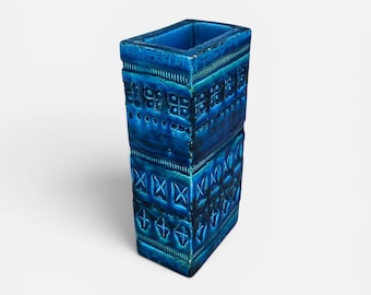 May include: A rectangular, ceramic vase in a vibrant shade of blue. The vase features a textured surface with geometric patterns, including squares, crosses, and horizontal lines. The open top reveals a hollow interior, suitable for displaying flowers or as a decorative piece.