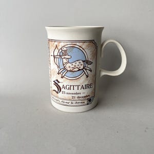 Op de afbeelding: Een witte keramische mok met een Boogschutter sterrenbeeld ontwerp. Het ontwerp bevat een centaur met pijl en boog, een blauwe achtergrond en het woord "SAGITTAIRE". De mok heeft een gebogen handvat.