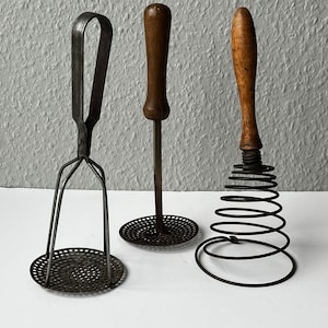 May include: Three vintage kitchen tools: a metal potato masher with a wire frame, a wooden handled potato ricer, and a metal spring-coil whisk with a wooden handle.