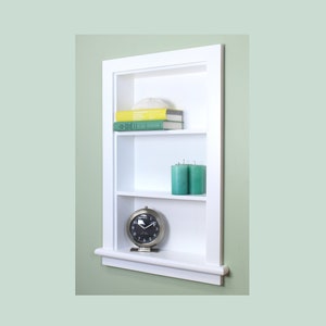 May include: A white, recessed wall shelf with three levels. The top shelf holds books and a hat, the middle shelf has two teal candles, and the bottom shelf displays a silver-toned clock. The shelf is mounted on a light green wall.