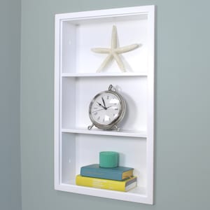 May include: White recessed shelf unit with three shelves. The top shelf has a white starfish, the middle shelf has a silver clock, and the bottom shelf has a stack of books and a teal candle.