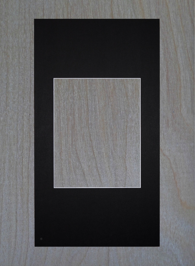 May include: A black picture frame with a white mat, creating a double border around a blank space in the center. The frame is on a light wood background.