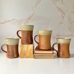 May include: Four ceramic mugs with a two-tone design featuring a speckled cream-colored upper section and a smooth, warm terracotta-colored lower body. Each mug has a curved handle and a slightly flared rim, arranged in a group.