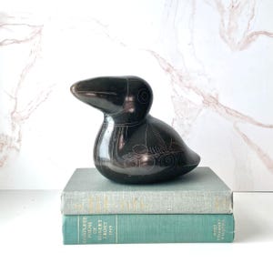 May include: A smooth, dark brown, bird-shaped sculpture featuring etched floral and leaf patterns on its side. The figurine has a rounded body and a prominent beak, resting on top of two stacked books, one light gray and one teal, with visible text.