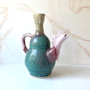 May include: A ceramic pitcher with a bulbous body glazed in deep green, featuring a tall neck with a light beige finish. The handle and spout are sculpted in a pale pink hue, with the spout shaped like an open-mouthed bird head.