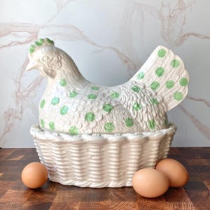 May include: A cream-colored ceramic hen-shaped lidded container with light green polka dots, resting on a base designed to resemble a woven basket. The hen features textured feather details and a green comb, accompanied by three brown eggs.
