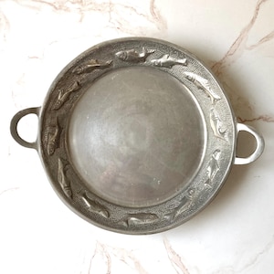 May include: A round silver-toned metal serving tray with two side handles. The wide rim features a textured background with a repeating embossed pattern of swimming fish circling the smooth, plain center of the circular platter.