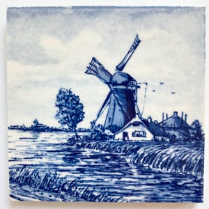 May include: Square ceramic tile featuring a blue and white illustration of a Dutch windmill scene. The artwork depicts a windmill, a house, a tree, and a body of water under a cloudy sky. The tile has a slightly aged appearance.