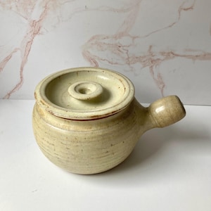 May include: A round, cream-colored ceramic pot with a matching lid and a single side handle. The surface features a speckled texture with subtle horizontal ridges and brown accents along the rim and handle, showcasing a handcrafted appearance.