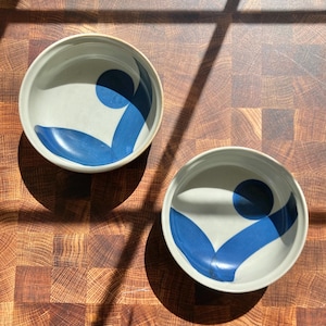 May include: Two ceramic bowls with a light gray interior and a blue abstract design. The design features a blue circle and two curved blue shapes. The bowls are on a wooden surface.