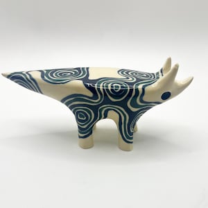 May include: A ceramic figurine of a four-legged animal with a pointed tail and two horns on its head. The off-white surface is decorated with dark blue swirling, organic patterns that cover the body, legs, and head, including a dark blue eye.