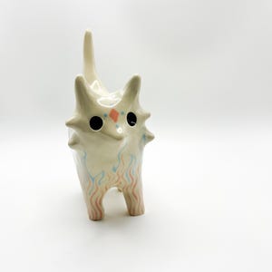 May include: A cream-colored ceramic animal figurine with a pointed tail and ears. The creature features large black circular eyes, a small orange diamond marking on the forehead, and delicate blue and orange flame-like patterns on its legs.